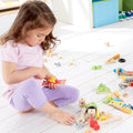 A young girl in a light purple outfit sits on the floor in a bright room, playing with colorful wooden building toys from her Hape Fix-It Tool Box scattered around her. She appears focused as she manipulates a yellow and red piece, surrounded by various bolts and blocks.