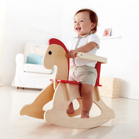 A cheerful toddler with light brown hair is sitting on the Hape Grow-with-me Rocking Horse. The child, dressed in a white shirt and beige shorts, is smiling while riding the Hape toddler rocking toy in a bright, minimalist room with light-colored furniture and decor.