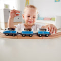 A young child with blonde hair is joyfully playing with a Hape Intercity Train on a curved wooden track. The background features a white-walled room adorned with child-friendly furniture, including a dresser and a bed. The child is smiling and raising one arm in excitement.