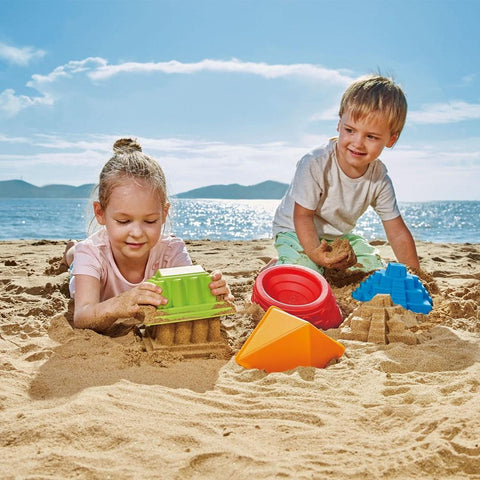 On a sunny beach, two children play in the sand surrounded by colorful beach toys. The girl on the left is focused on building with a Hape Mayan Pyramid mold, crafting small sand temples, while the boy on the right, wearing a white shirt, smiles and watches as he sifts through the sand. The ocean and distant hills can be seen in the background.