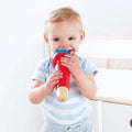 A smiling toddler with blonde hair, dressed in a blue and white striped shirt, holds a Hape Mighty Echo Microphone in red and blue. The child seems to be pretending to sing into the microphone against the backdrop of a light-colored room featuring a white wall and some furniture. This scene makes for an entertaining gift idea from Hape's musical instrument collection.