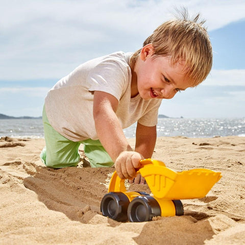 A young boy with blonde hair, dressed in a white t-shirt and green pants, is playing on a sandy beach with his Hape Monster Digger. The ocean and a distant coastline can be seen in the background under a clear blue sky.