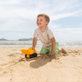 A young child in a white t-shirt and light green shorts is playing with a yellow Hape Monster Digger beach toy dump truck on a sandy beach. The child is smiling and kneeling in the sand. The ocean and distant hills are visible in the background under a partly cloudy sky.