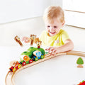 A smiling little boy with blond hair is playing with the Hape Music and Monkeys Railway, embarking on an animal adventure. The train set, vibrant with colorful animal figures including a monkey and an elephant, is arranged on a white table, complemented by a nearby small green tree.