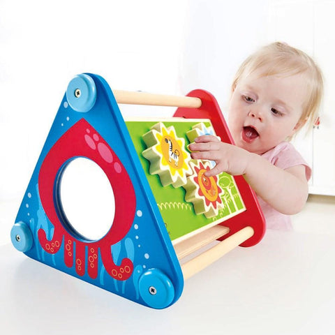 A young child interacts with the Hape Take-Along Activity Box. The toy, designed by Hape, features a triangular shape with a blue and red base, a circular mirror on one side, and colorful spinning gears on the green side panel. The child is touching the gears while looking at the toy with curiosity, enhancing their fine motor skills.