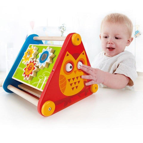 A toddler enjoys playing with the Hape Take-Along Activity Box, a vibrant and engaging wooden toy from Hape, featuring a triangular shape and an owl design on the side. The activity box includes rotating gears along with smiling sun and flower illustrations, captivating the child's curiosity and enhancing fine motor skills as they interact with its various elements.