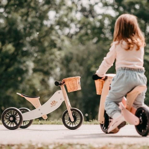 A young child with long hair rides a Kinderboard Kinderfeet Tiny Tot Plus - White, a wooden balance bike, on a paved path in a park. Another 2-in-1 bike with a wicker basket at the front is parked nearby. The scene is surrounded by green trees and foliage, creating a peaceful outdoor setting.