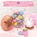 A clear jar filled with colorful bath bombs from the KLUTZ Make Your Own Bath Bombs Scented Bakery set sits on a pink surface beside a white, bunny-shaped dispenser. The jar has a copper lid resting nearby. A pink ribbon lays in the background, and text above reads, "Make up to 12 bath bombs for some fizzy fun!" Perfect as a gift box surprise!