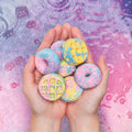 Hands holding a selection of small, colorful Klutz Make Your Own Bath Bombs Scented Bakery by KLUTZ in various shapes, including donuts and waffles. The bath bombs are made with baking soda and come in pastel colors of blue, pink, yellow, and purple against a vibrant, abstract background in similar shades. Perfect for a delightful gift box!