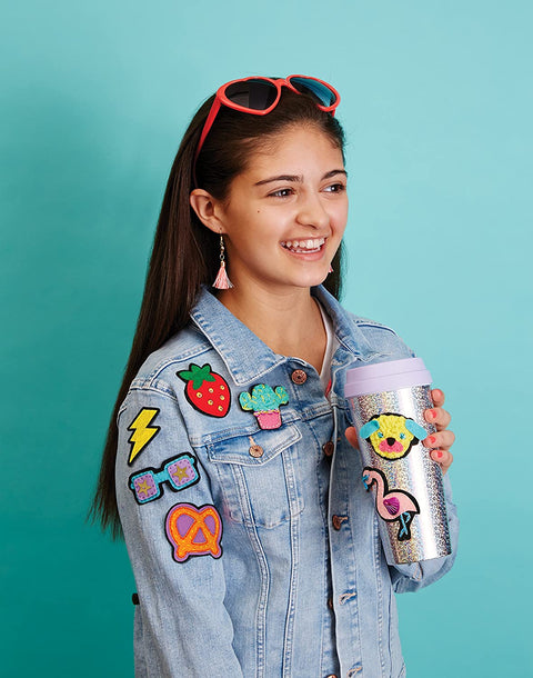 A young person with long brown hair and red sunglasses perched on their head smiles while holding a glittery cup. They are wearing a denim jacket adorned with colorful stick-on patches from the "Klutz Make Your Own Stick on Patches" kit by KLUTZ, which include designs like a strawberry, lightning bolt, flamingo, pretzel, and drink cup. The background is turquoise.