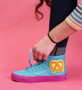 A person with long, curly hair ties the laces of a bright blue high-top sneaker with a vibrant pink sole. Adorning the side of the sneaker is a large, decorative patch featuring a pretzel, made using Klutz Make Your Own Stick On Patches from KLUTZ. The background is pink, and the person is dressed in dark skinny jeans and a striped shirt.