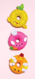 Three plushies from the KLUTZ Sew Your Own Donut Animals kit, each designed like donut toys with cute, embroidered faces, are arranged vertically on a pink background. The yellow one resembles a pineapple with green leaves, the pink one features white swirls and green leaves, and the orange one is styled like an orange with white flowers.