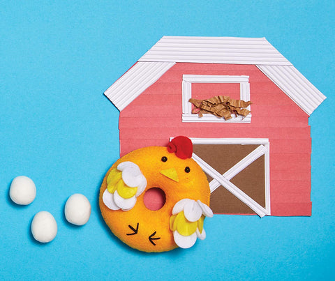 A whimsical scene highlights a felt donut from the Klutz Sew Your Own Donut Animals kit by KLUTZ, styled as a chicken complete with wings and a red comb. The donut is displayed on a blue surface with a pink and white paper barn in the background, accompanied by three white eggs placed to its left.