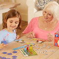 A young girl and an elderly woman enjoy playing the Orchard Toys Numbers Bear Game together at a wooden table. The girl holds a game piece from the Orchard Toys brand while the woman smiles, engaging in solving mathematical problems. The table is scattered with game pieces and cards.