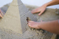 Hands working on a QUUT Pira Stackable Pyramid, carefully smoothing out the surface and adding detailed features. The pyramid, complete with an archway at its base that resembles an entrance, echoes the look of intricate architectural sculptures. Sand is spread around the base of the QUUT pyramid on a flat surface.