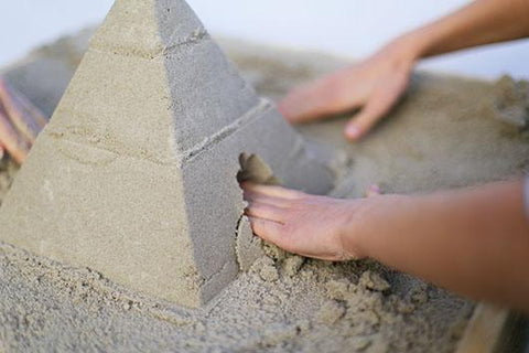 Hands working on a QUUT Pira Stackable Pyramid, carefully smoothing out the surface and adding detailed features. The pyramid, complete with an archway at its base that resembles an entrance, echoes the look of intricate architectural sculptures. Sand is spread around the base of the QUUT pyramid on a flat surface.