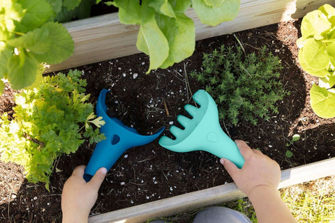 Two hands holding QUUT Raki Rake & Digger - Pink, crafted by QUUT, work in a raised garden bed. The bed has small plants growing in soil, surrounded by leafy greens and herbs. The vibrant pink tools contrast with the earthy tones of the garden soil, making gardening fun for all ages.