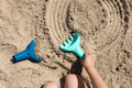 A person rakes the sand with a claw-shaped children's garden tool from QUUT, known as the QUUT Raki Rake & Digger in pink, creating a circular pattern. Nearby, a blue shovel lies on the sand. Knees are partially visible at the bottom of the image, suggesting a playful sand activity with an open-ended toy.