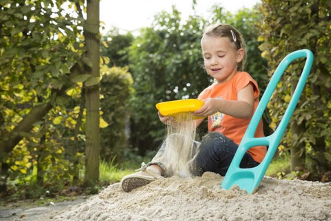 A young girl, sitting in a sandbox outdoors, pours sand with the blue QUUT Scoppi Spade by QUUT. She is wearing an orange shirt, jeans, and sneakers. To her right, there is a blue plastic slide and a kids beach toy sand sifter. The background features green shrubs and a sunny sky.