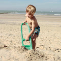 A young boy in swim trunks digs in the sandy beach with a large blue QUUT Scoppi Spade, an iconic kids beach toy from QUUT, under a clear sky. The beach and sea stretch out behind him with a few sailboats visible in the distance.