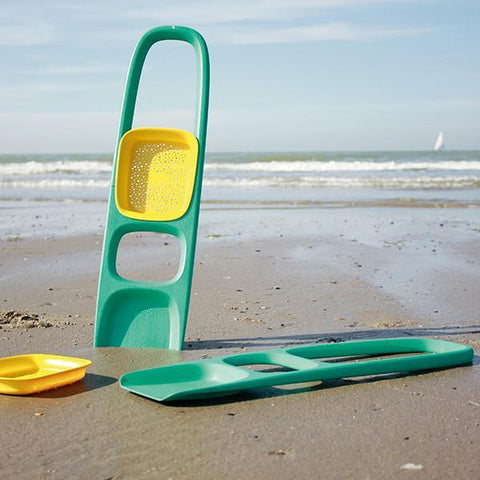 A cherry red QUUT Scoppi Spade from the brand QUUT, featuring a detachable sand sifter, is placed in the sand near the shoreline. The ocean waves gently roll in the background under a clear blue sky, with a sailboat visible in the distance.