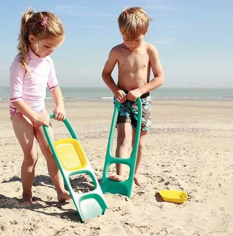 Two children are playing on a sandy beach with toy sieves and shovels. The girl, dressed in a pink rash guard, and the boy, wearing swim trunks, are engaged in scooping sand with their QUUT Scoppi Spade - Cherry Red on a sunny day. The ocean and blue sky are visible in the background.