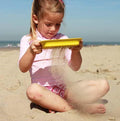 A young girl wearing a pink rash guard and patterned shorts is sitting on the sandy beach, sifting sand through her yellow kids beach toy. The QUUT Scoppi Spade in Cherry Red, with its detachable sand sifter design by QUUT, makes playtime even more fun. The sky is clear, and the ocean is visible in the background.