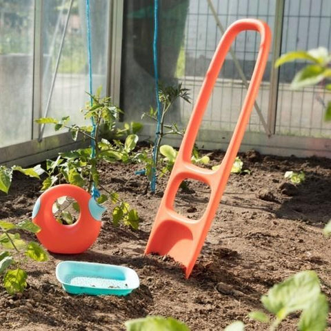 A garden scene with young plant seedlings growing in soil. In the foreground, there are three bright-colored gardening tools: an orange and blue scoop, an orange digging tool, and a cherry red QUUT Scoppi Spade with a detachable sand sifter from the QUUT design collection, all placed near the plants.