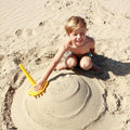 A young boy wearing swim trunks is building a sandcastle with a green QUUT Triplet Beach & Bath Toy on a sandy beach. He is smiling and appears to be excitedly engaged in his activity. The sandcastle is shaped with concentric circles.