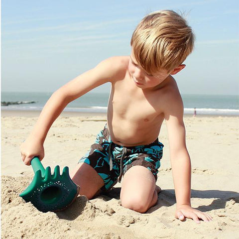 A young boy with blond hair is digging at the beach using a QUUT Triplet Beach & Bath Toy in green. He is shirtless and wearing dark swim trunks with blue patterns. The ocean and a clear sky can be seen in the background.