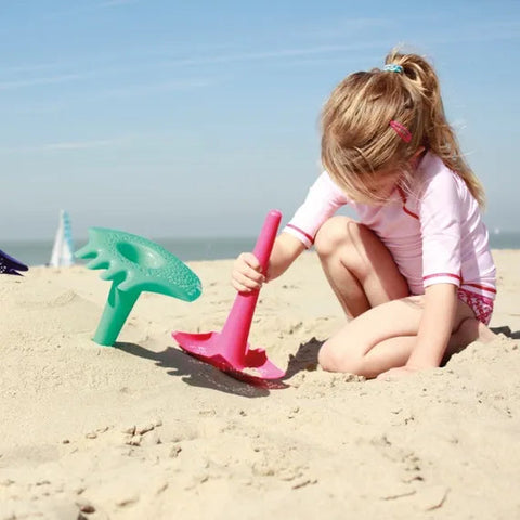 A young girl with blond hair tied in a ponytail is sitting on a sandy beach. Wearing a white and pink rash guard and shorts, she plays with a red, multifunctional sand tool. Nearby, the QUUT Triplet Beach & Bath Toy in green from QUUT is stuck in the sand. The sky above is clear and blue.