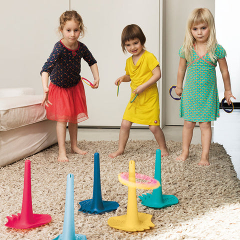 Three young children stand barefoot on a cream-colored rug, playing a ring toss game in a living room. The children hold colorful QUUT Triplet Beach & Bath Toys - Green and aim to toss them onto tall, cone-shaped stands of various colors. A light sofa sits to the left and a white wall is in the background. Their lively activity brings the fun spirit of a beach day indoors, courtesy of QUUT.