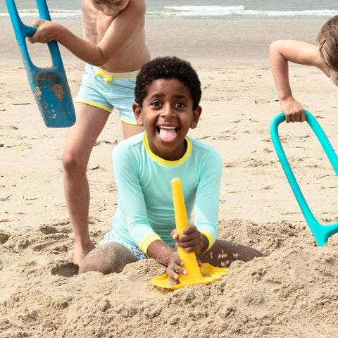 Three children play on the beach. The child in the center, wearing a light blue swimsuit, sits in the sand holding a yellow QUUT Triplet Beach & Bath Toy - Green and smiling with their tongue out. The other two children, also in swimsuits, use blue shovels to dig around them.