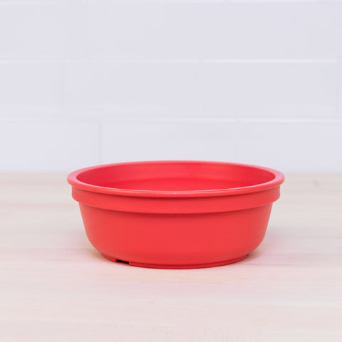 A REPLAY Re-Play Bowl, distinguished by its round, red design and slightly raised sides, rests on a light wooden surface. The backdrop is a clean white tiled wall. This colorful piece of tableware is crafted from recycled plastic bowls, ensuring it is both safe and suitable for kids.