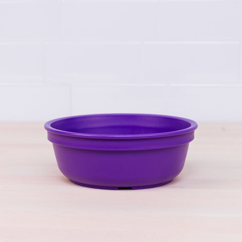 A vibrant tableware set, featuring a Re-Play Bowl from REPLAY, made from recycled purple plastic and round in shape, sits on a light wooden surface with a white tiled wall in the background.