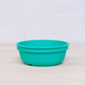 A Re-Play Bowl from REPLAY, characterized by its turquoise color and shallow design, is part of a vibrant tableware set and sits on a light wooden surface with a white tile wall in the background. The bowl features a smooth, rounded design and an even matte finish. Made from recycled plastic, this bowl is safe for kids.