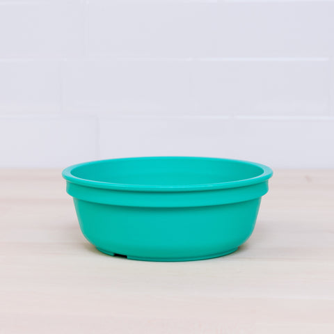 A Re-Play Bowl from REPLAY, characterized by its turquoise color and shallow design, is part of a vibrant tableware set and sits on a light wooden surface with a white tile wall in the background. The bowl features a smooth, rounded design and an even matte finish. Made from recycled plastic, this bowl is safe for kids.