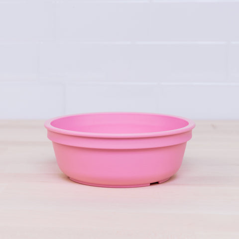 A vibrant pink Re-Play Bowl from REPLAY's colorful tableware set is placed on a light wooden surface with a white tiled wall in the background. Made from recycled plastic, the bowl is empty and features a simple, smooth design that is safe for kids.