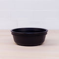 A REPLAY Re-Play Bowl, black and round, made from recycled plastic sits empty on a light wooden surface with a white tiled wall background.