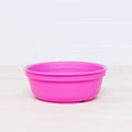 A Re-Play Bowl by REPLAY, made from recycled plastic and featuring a smooth surface with a slightly rounded rim for a simple and functional appearance that's safe for kids, sits on a light wooden surface with a white tiled background. This bright pink plastic bowl is part of a vibrant tableware set.