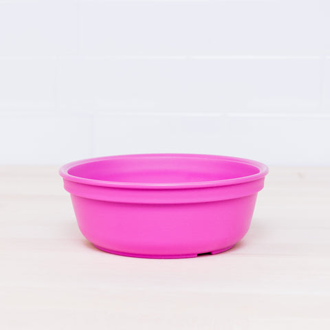 A Re-Play Bowl by REPLAY, made from recycled plastic and featuring a smooth surface with a slightly rounded rim for a simple and functional appearance that's safe for kids, sits on a light wooden surface with a white tiled background. This bright pink plastic bowl is part of a vibrant tableware set.