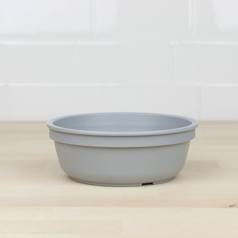 A Re-Play Bowl from REPLAY, made of gray recycled plastic, is placed on a light wood surface. The background consists of white tiled walls. The bowl has a simple, unadorned design, ideal and safe for kids.