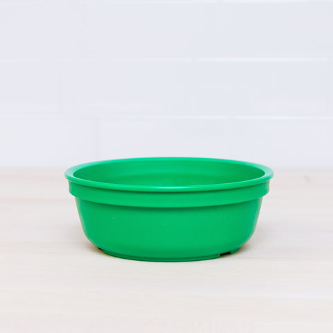 The Re-Play Bowl by REPLAY, crafted from recycled green plastic and featuring a round shape, rests on a light wooden surface with a white tiled wall in the background. Safe for kids, the bowl's simple design stands out against its plain surroundings.