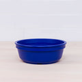 A Re-Play Bowl from REPLAY, featuring a round, solid blue plastic design, sits on a light wooden surface with a white tiled wall in the background. This vibrant piece from a colorful tableware set is made from recycled plastic and is safe for kids. The bowl boasts a simple, smooth design and is currently empty.