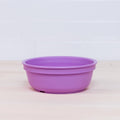 A vibrant Re-Play Bowl from the REPLAY kid-safe tableware set is placed on a light wooden surface against a white tiled wall. The purple bowl boasts a simple, design-free aesthetic and is made from recycled plastic.