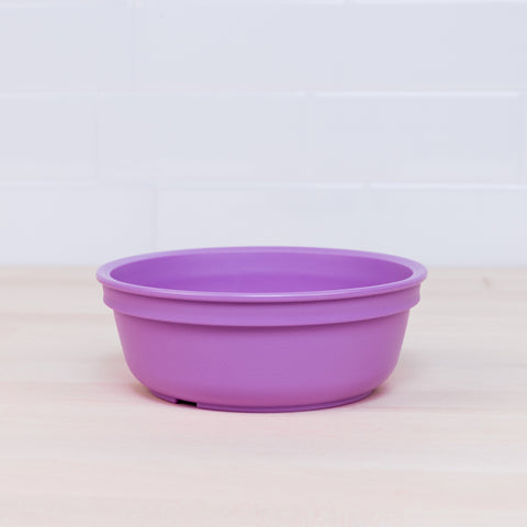 A vibrant Re-Play Bowl from the REPLAY kid-safe tableware set is placed on a light wooden surface against a white tiled wall. The purple bowl boasts a simple, design-free aesthetic and is made from recycled plastic.