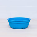 A Re-Play Bowl in blue, part of the vibrant REPLAY tableware set, sits on a light wooden surface with a white tiled background. Made from recycled plastic, this round bowl features a smooth finish and a slightly raised rim, making it safe for kids.