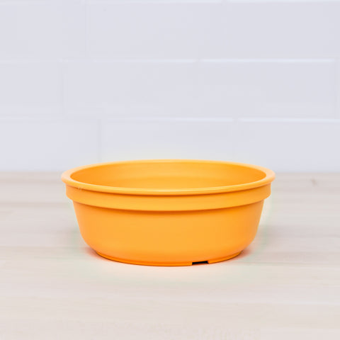 A Re-Play Bowl by REPLAY, crafted from recycled vibrant orange plastic with a round shape and slightly flared rim, is placed on a light wooden surface. Safe for kids, it seamlessly blends into any home with its cheerful design against the white tiled background.