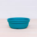 A Re-Play Bowl from REPLAY, part of a vibrant tableware set, sits on a light wooden surface with a white tiled wall in the background. Made from recycled plastic, this teal bowl features a simple, smooth design that is safe for kids.