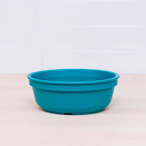 A Re-Play Bowl from REPLAY, part of a vibrant tableware set, sits on a light wooden surface with a white tiled wall in the background. Made from recycled plastic, this teal bowl features a simple, smooth design that is safe for kids.